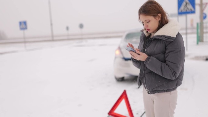 一名年轻的女司机在冬天的雪地里，在湿滑的道路上撞坏了自己的车，她在车旁边呼叫了一辆带有紧急三角标志的