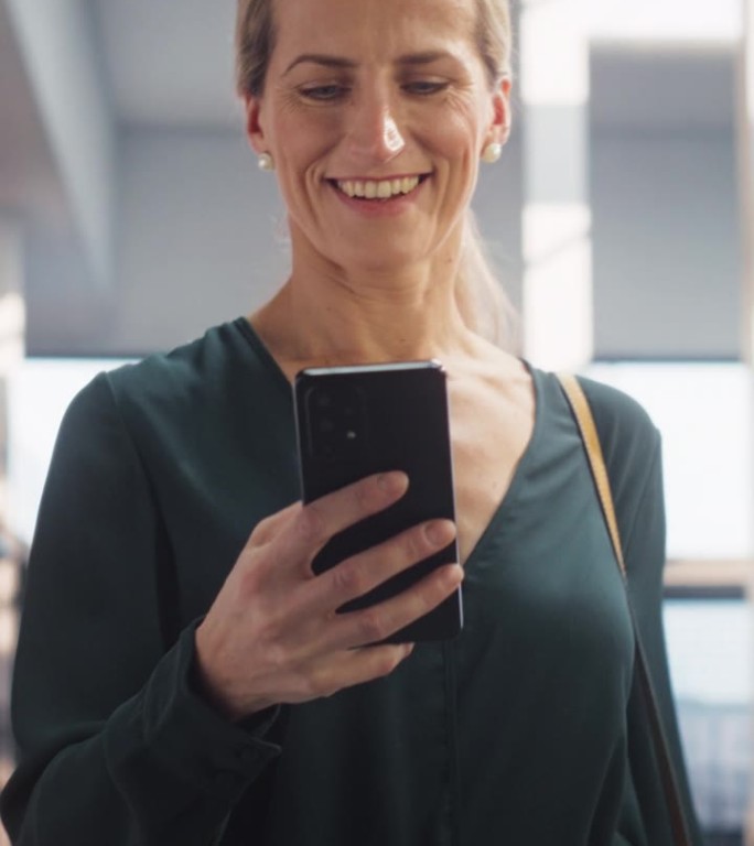 垂直屏幕:一个快乐的白人女性在公司办公室用智能手机发短信，微笑着走着。市场经理检查社交媒体趋势并保持