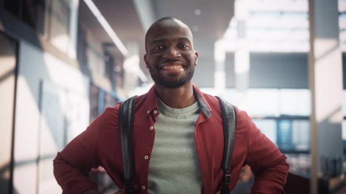 一个快乐的男学生站在现代公共图书馆的肖像。年轻的黑人男子微笑着看着镜头。学生背着书包在大学里摆姿势。