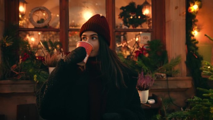 穿着毛皮大衣，戴着帽子，戴着连指手套的美丽开朗的女孩在圣诞市场或集市上，在节日花环附近喝着热饮，享受