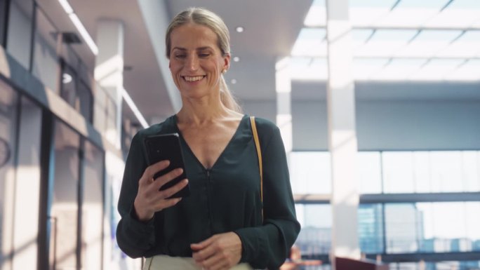 一个快乐的白人女性在她的智能手机上发短信，微笑着走在公司办公室。女性高级营销经理查看社交媒体趋势并保