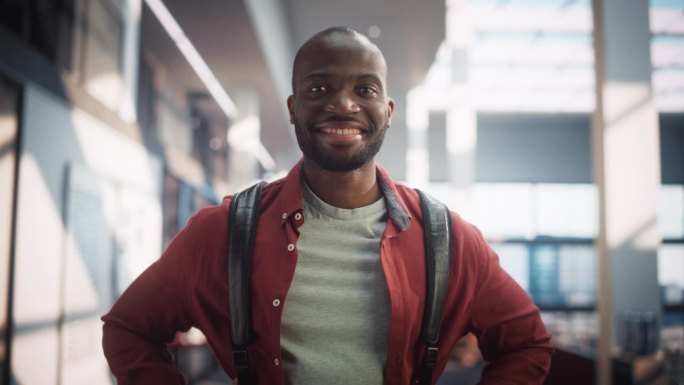 一个快乐的男学生站在现代公共图书馆的肖像。年轻的黑人男子微笑着看着镜头。学生背着书包在大学里摆姿势。