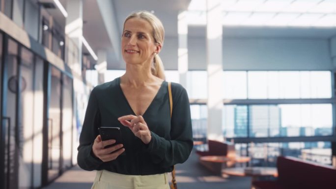 白人女项目经理走在现代办公楼走廊上，用智能手机看大城市。在国际电子商务公司工作的职业女性