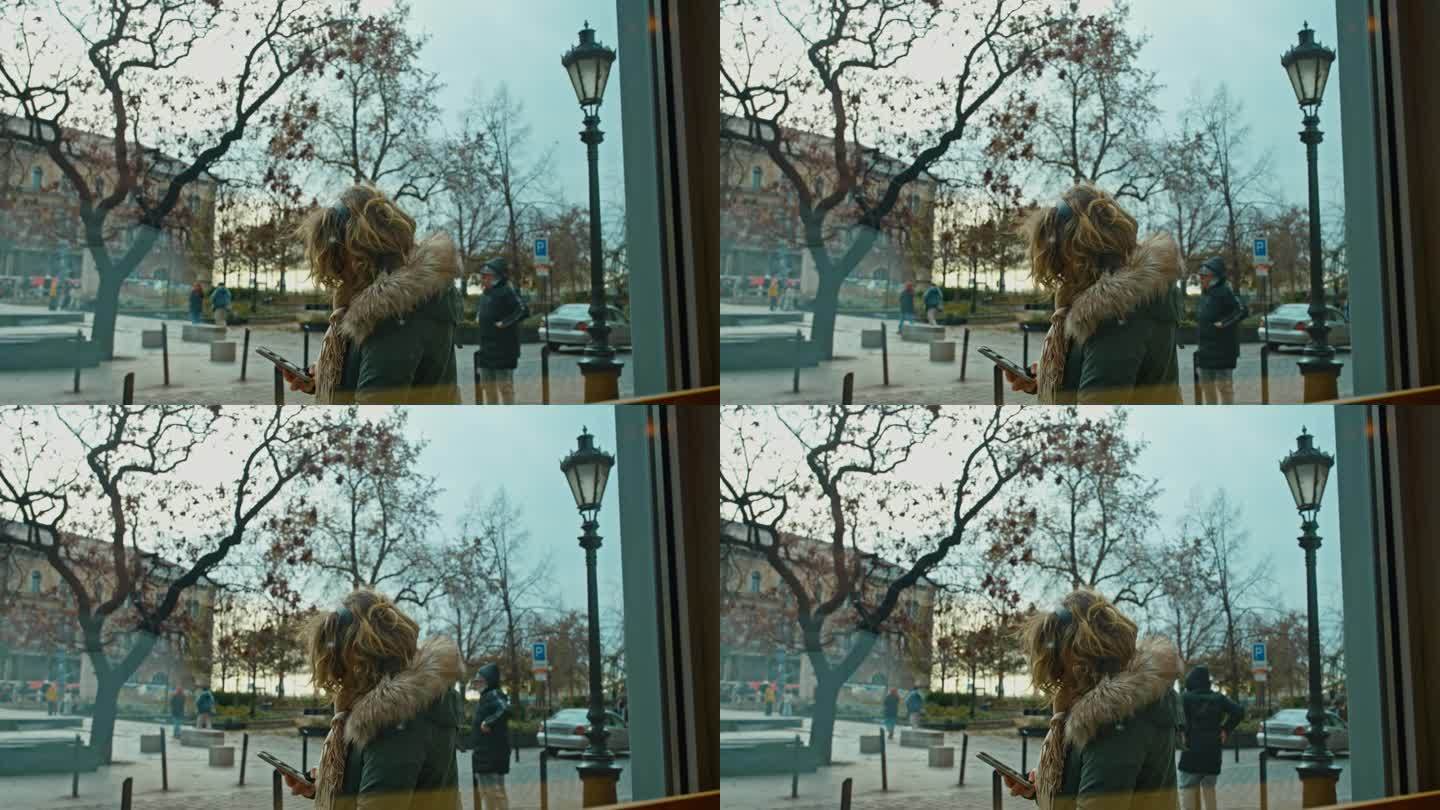 匈牙利布达佩斯，透过玻璃窗看到的穿冬衣的女人在城市街道上使用智能手机的侧视图