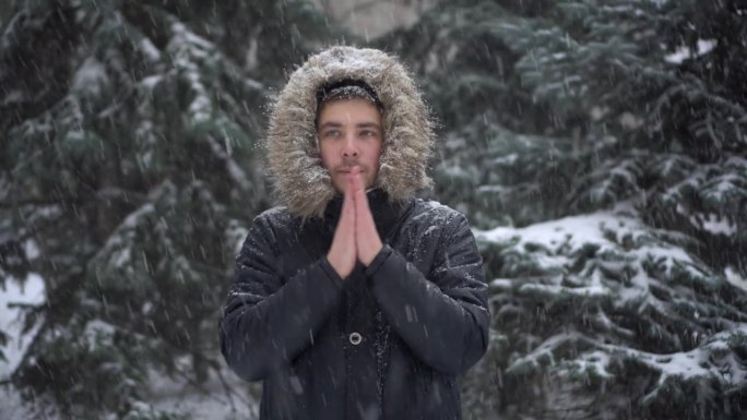 大雪下，一个年轻人站在冷杉树的背景下，用手搓着御寒。一名身穿羽绒服、戴着兜帽的男子站在那里看着镜头。