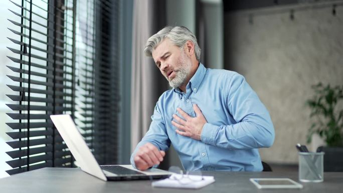 一位白发苍苍、大胡子的成熟商人在办公室用笔记本电脑工作时突发心脏病。