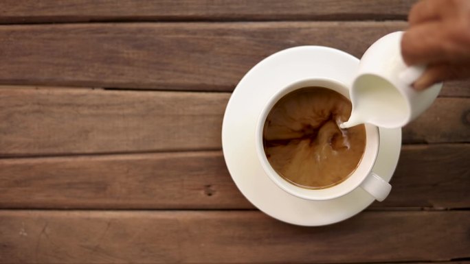 将牛奶倒入黑咖啡中，倒进空的白色杯中，杯底是木制的茶托。