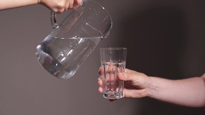 一只手拿着一个透明的玻璃杯，透明的干净的饮用水从灰色背景的滗水器中倒进去。饮用水。水标准。纯水