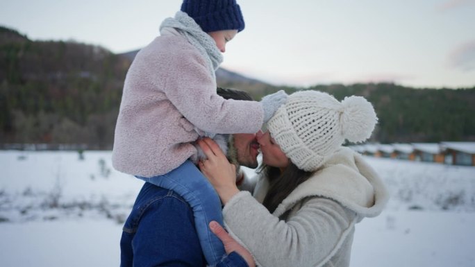 快乐的年轻家庭在冬季假期与小孩子在雪地里接吻。