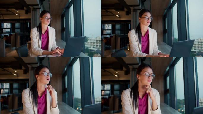 一个年轻的黑发女孩，戴着圆眼镜，穿着白色夹克和粉色衬衫，坐在办公室的全景窗旁，用笔记本电脑工作