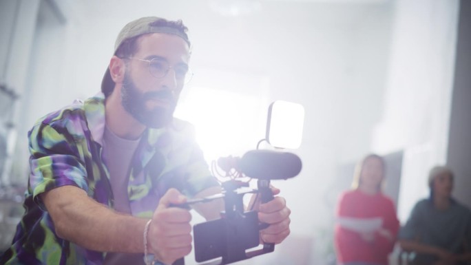 热情的男性摄像师的肖像拍摄项目使用智能手机钻机设备在一个明亮的工作室。专业的在线内容制作者在拍摄时指