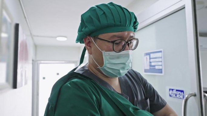 图为做完手术的医生脱下手术帽，助手帮他脱下绿色手术服，走在办公室前的走廊上。