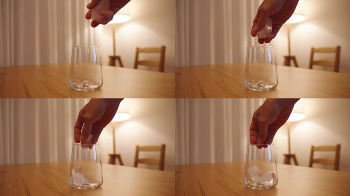 一只女性的手把冰块扔进木桌上的空杯子里。白色房间里昏暗的灯光，背景是落地灯。
