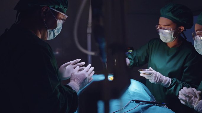 一名男外科医生和他的助手密切注视着，一名护士用镊子拿着棉球，在病人腹部的切口上涂聚维酮碘液消毒。