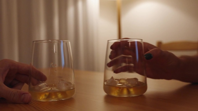 男人和女人把他们的威士忌酒杯并排放在一张木桌上。这个场景是在一个白色的房间里被昏暗而温暖的灯光照亮的