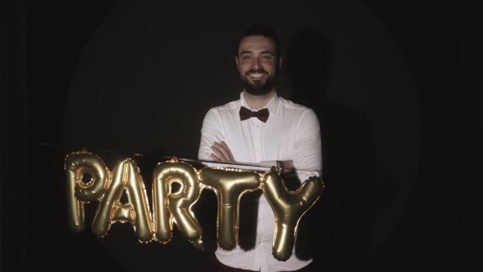 一名男子双臂交叉，对着镜头微笑，而“派对”气球出现在他面前