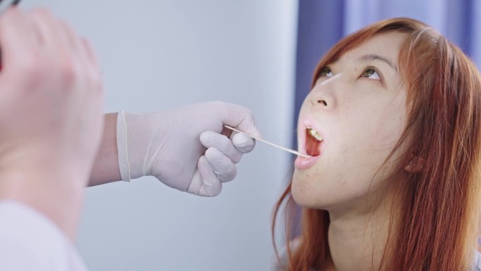 在医学检查室里，一名男性耳鼻喉科医生正在用压舌板和手电筒检查她的喉咙，一名女病人张开嘴的特写。