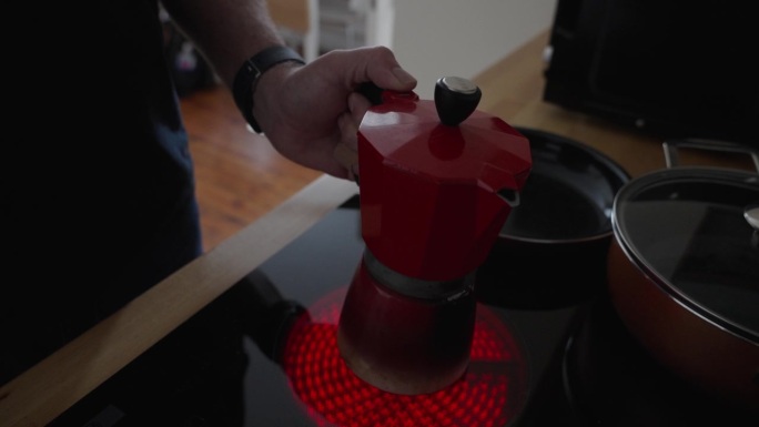 一个人打开陶瓷电炉的燃烧器，把一个红色的浓缩咖啡壶放在灼热的发光元件上