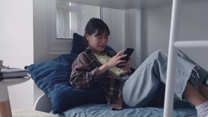 一名大学生正在宿舍舒适的卧室里用智能手机在社交网络上聊天。