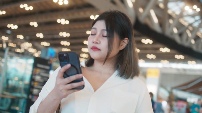 积极的年轻亚洲女性穿着舒适的衣服，使用智能手机，查看航班信息板，在国际机场行走和站立，快乐的女乘客拿