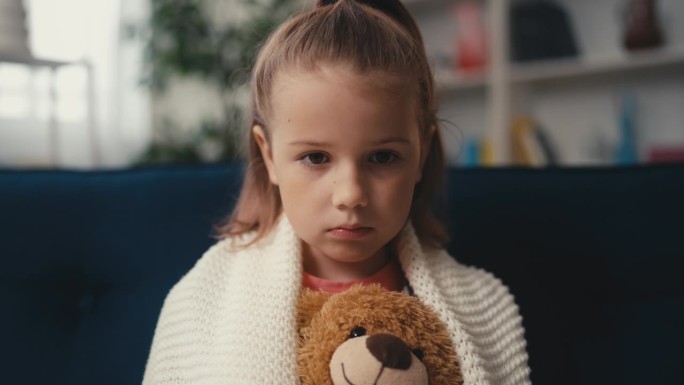 不开心的小女孩抱着泰迪熊独自坐在沙发上，悲伤