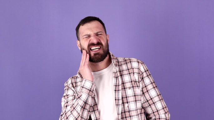 年轻的大胡子男子的肖像与牙齿疼痛的问题在口腔孤立的紫色背景在工作室。