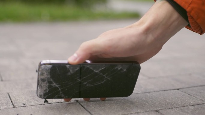 手机摔在人行道上，一名男子拿着手机捡了起来，手里拿着一个坏了屏幕的智能手机