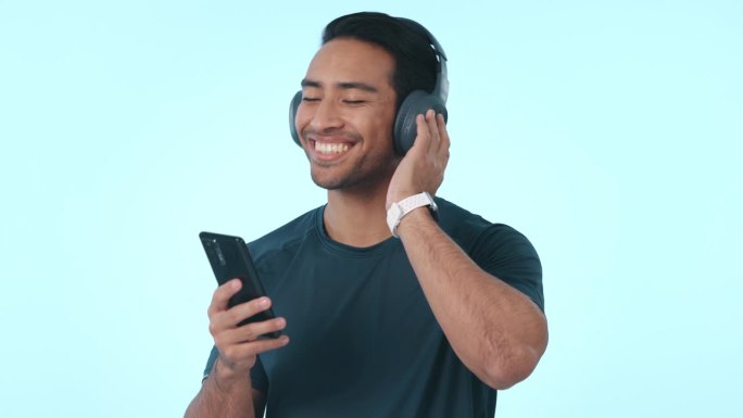 快乐的亚洲人，手机和耳机听音乐或播客在工作室的背景下。男士带着耳机微笑，在智能手机或应用程序上享受音