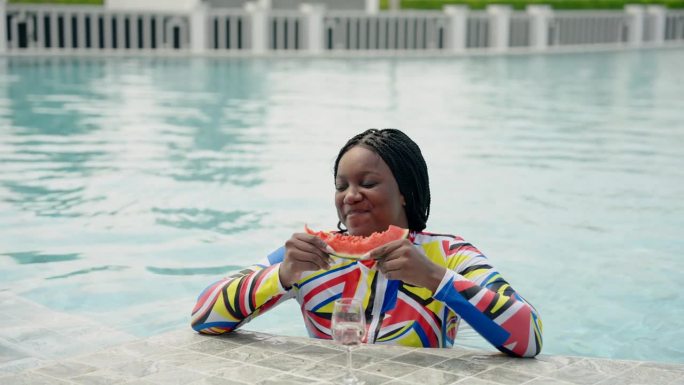 美丽的非洲女孩站在水里快乐地吃着西瓜在池塘里晒太阳在游泳池里戴着帽子和太阳镜。在一个晴朗的夏日周末一