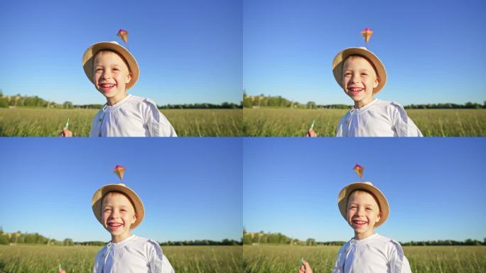 戴草帽的高加索男婴手持风筝在绿地上的特写肖像。孩子开心，龇牙咧嘴，哈哈大笑。夏季户外散步的休闲活动。