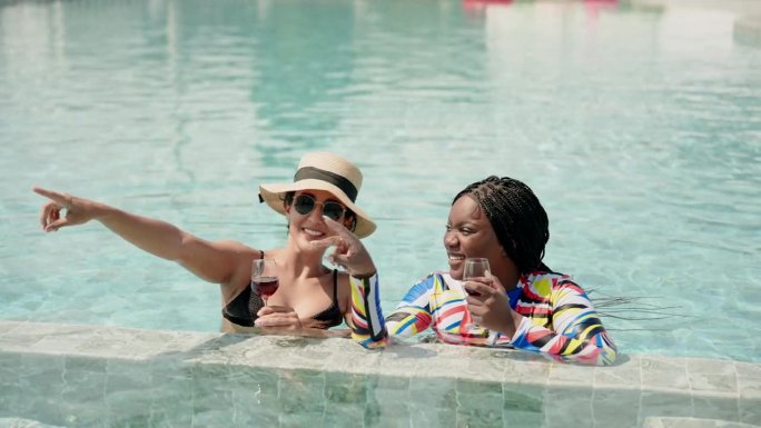 非洲女人和西班牙人，亲密的朋友，两个多民族的人站在水里喝着酒，愉快地聊天。在炎热的晴天里，在游泳池里