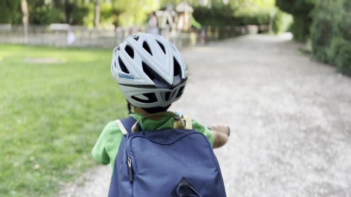 一个小男孩带着头盔和背包在安静空旷的街道上骑行，路过树木和操场