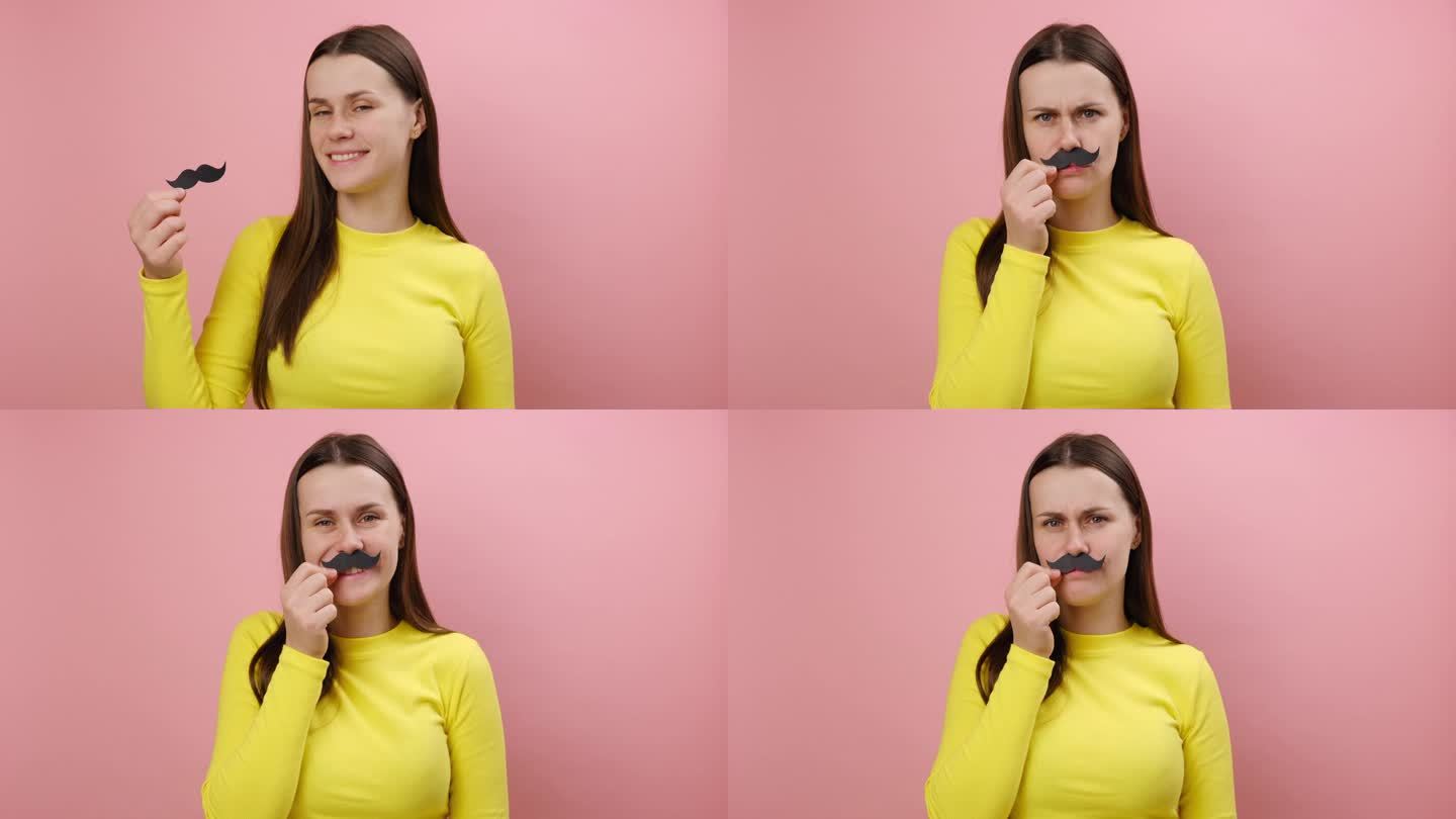 有趣俏皮的年轻白人女子留着假纸胡子，开心地看着相机，有节日的心情，穿着黄色毛衣，在工作室的纯粉色背景