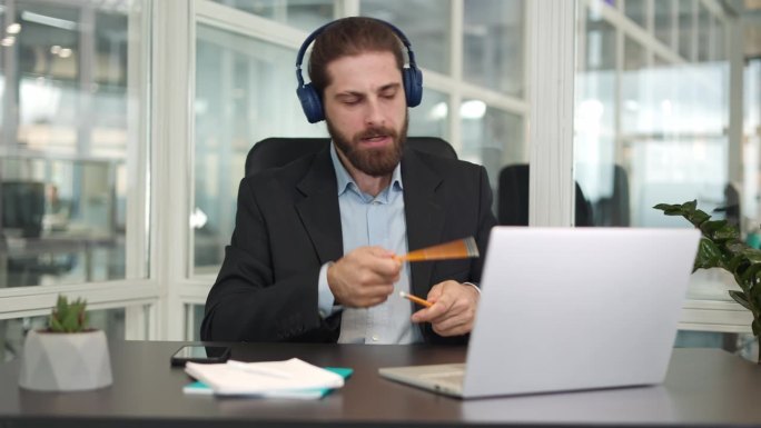 微笑的高加索商人用无线耳机听音乐，耳机连接到个人橱柜里的现代笔记本电脑。快乐的男性躁狂症患者用铅笔当