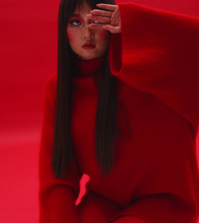 一个美丽的女孩穿着红色的衣服，在红色的背景下坐在摄影师面前摆姿势。垂直视频