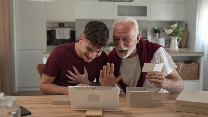 老人爷爷养老金领取者花白的头发和胡子与他的孙子十几岁的男孩收到礼物在盒子里打开阅读卡片快乐的微笑在家