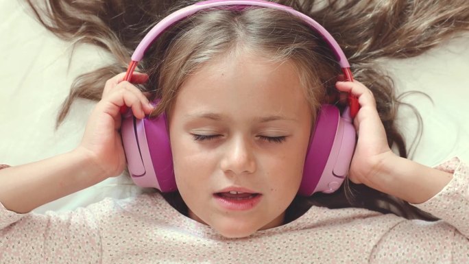 一个小女孩躺在卧室的床上，闭着眼睛戴着白色耳机听音乐。女孩手拿耳机，在家里动着头唱歌