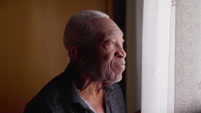 一位沉思的巴西黑人老人站在窗前，若有所思地望着窗外。非裔美国人的特写侧面脸从家里盯着外面