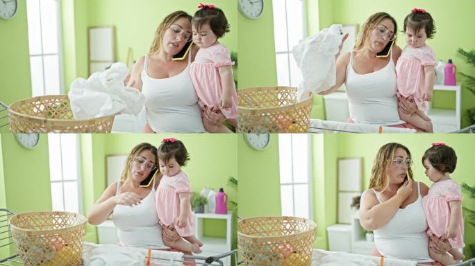 可爱的妈妈抱着她的宝贝女儿，一边讲着手机，一边悠闲地把衣服挂在晾衣绳上，享受着家里洗衣房的日常琐事