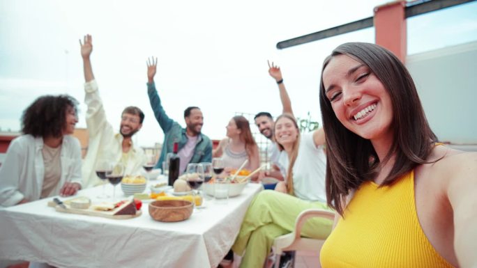 在前景中，一位年轻的成年女子和她的多种族朋友在家里屋顶的周末午餐派对上开心地笑着自拍。户外社交娱乐
