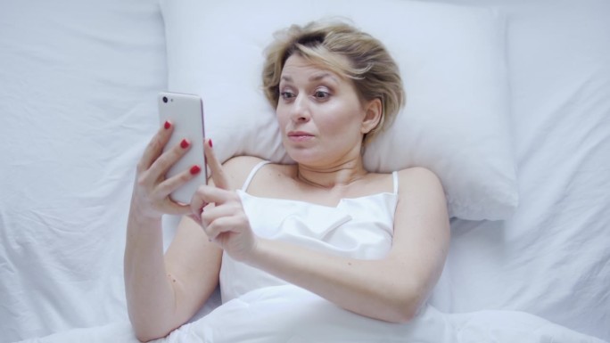 心情激动的女人躺在舒适的床上，用智能手机看最喜欢的电视剧或最喜欢的乐队的在线演唱会，睡眠问题