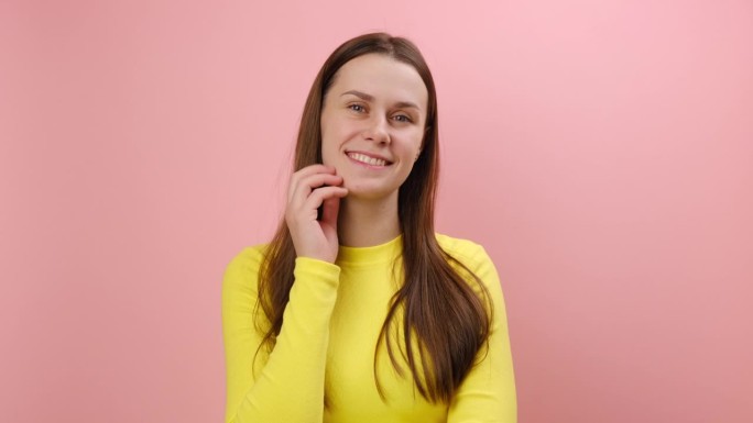 积极迷人的年轻白人女性的肖像，看着镜头，调情的表情，表达快乐的情绪，穿着黄色毛衣，在工作室粉红色的背