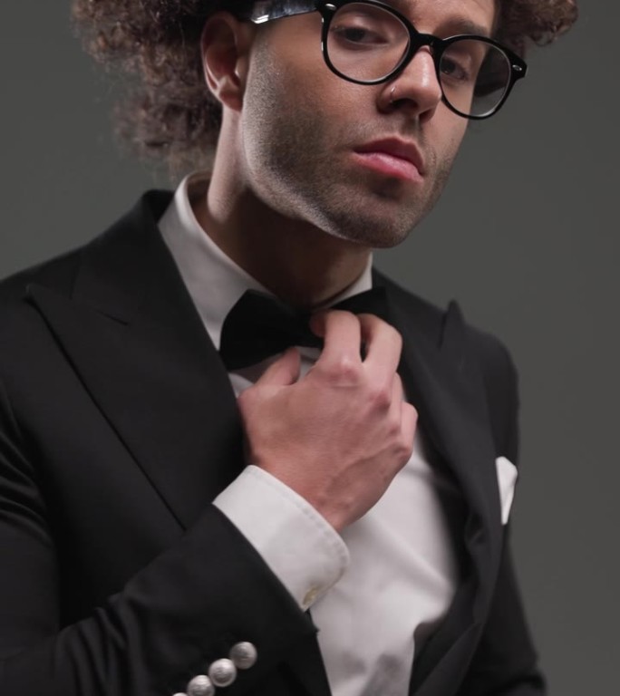 优雅的年轻人戴着眼镜看向一边，搓着手掌，在灰色的背景前调整着黑色领结