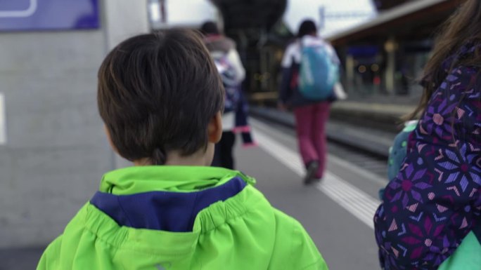 孩子的背影在火车月台上走向城市，旁边是他的兄弟姐妹。小男孩穿着雨衣和家人一起向前走