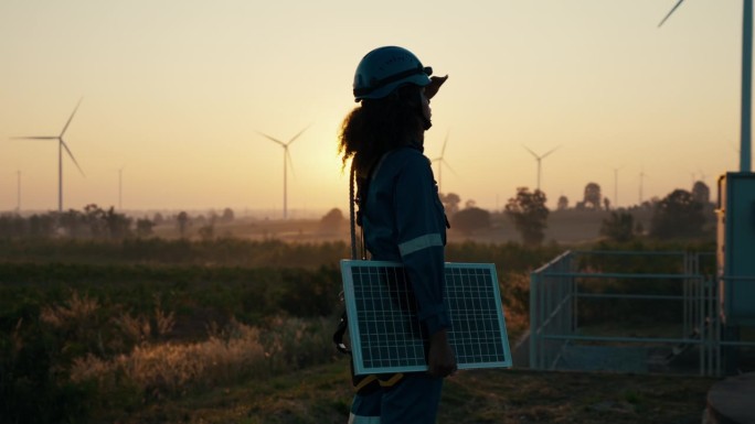 一名非洲女工程师手持太阳能电池板，勘察风力涡轮机发电场。