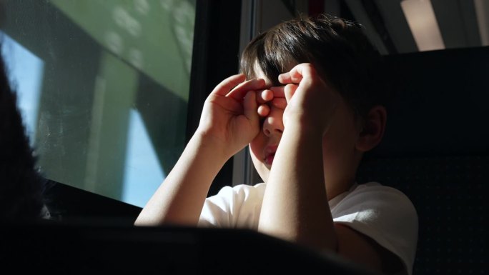 疲惫的孩子在行驶的火车上揉眼睛和脸，乘客孩子在窗户边打盹醒来，感觉很累