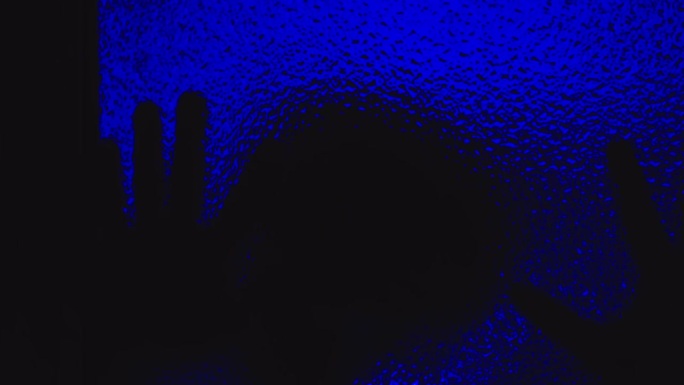 令人毛骨悚然的附身男子的剪影在令人难以忘怀的蓝光下在黑暗中爬在玻璃上