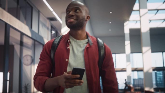 一个大学生的肖像，一边看手机，一边微笑着走向教室。年轻的黑人男子穿过大楼的大厅。低角度