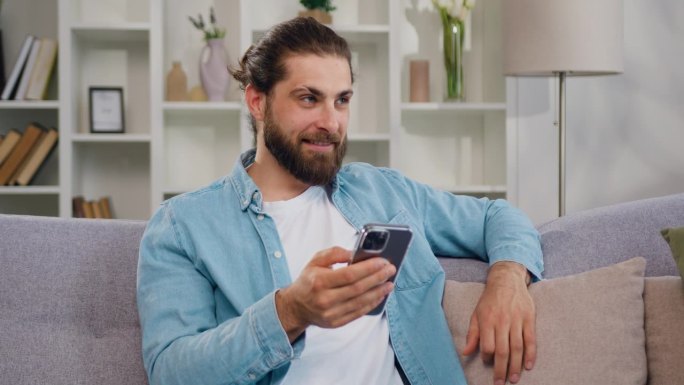 微笑的男子坐在时尚舒适的客厅的沙发沙发上，用智能手机远程在线工作。虚拟家庭办公，远程工作。自由职业者