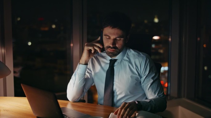 一个工作狂坐在办公桌旁讲电话，笔记本电脑近在咫尺。